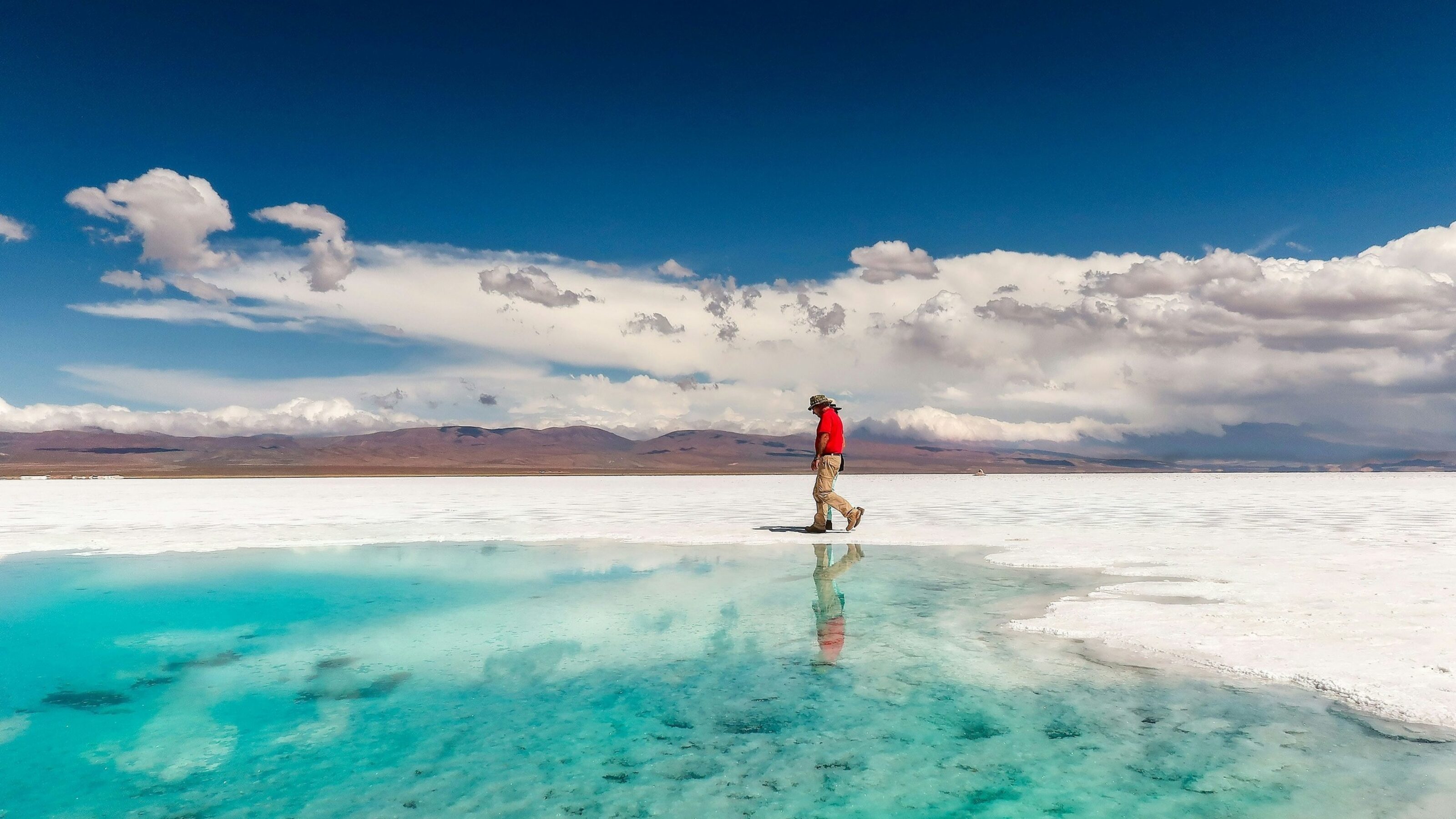 Person walking near a bright turquoise water pool on white salt flats with mountain reflections under a blue sky.
