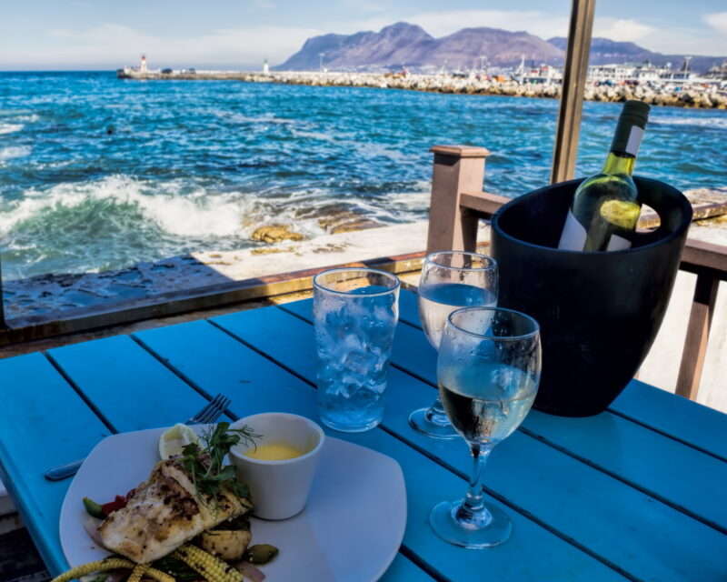 Grilled fish dish and white wine on a blue wooden table at a seaside restaurant with ocean waves in the background.