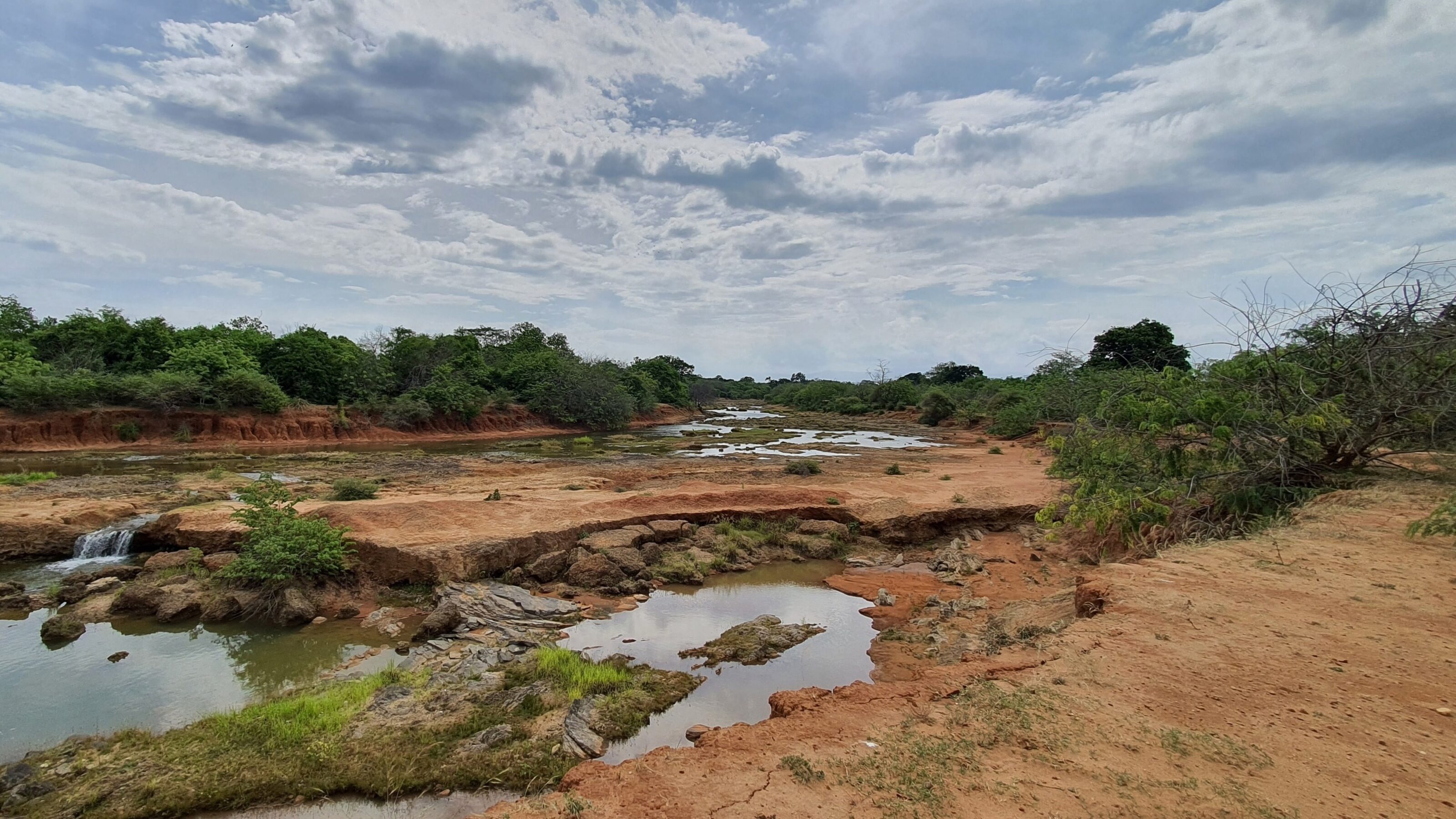 A rocky riverbed with small pools of water surrounded by red soil and green bushes under a vast sky.