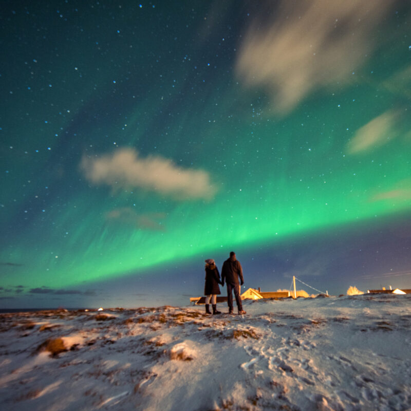 Couple in winter gear stands on a snowy hill under a bright green aurora borealis and a starry night sky.