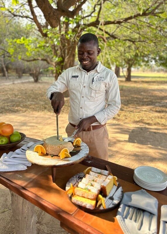 A man in a tan uniform slices a cake on a wooden table outdoors, surrounded by plates of sliced bread and fruit.