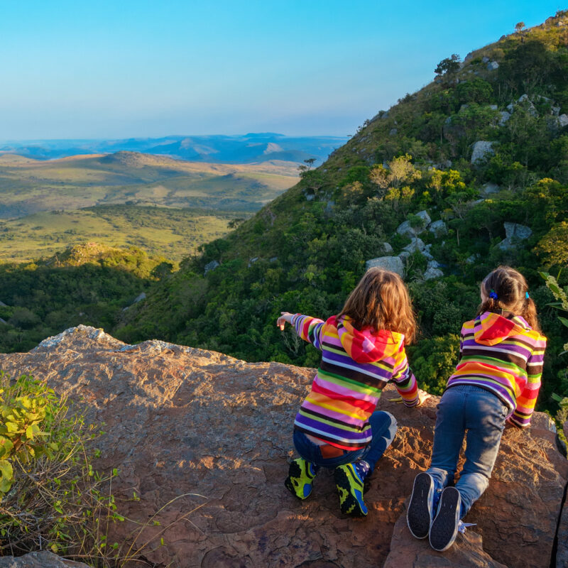 Two children in colorful hoodies sit on a rocky cliff edge, pointing toward a lush mountain valley landscape.