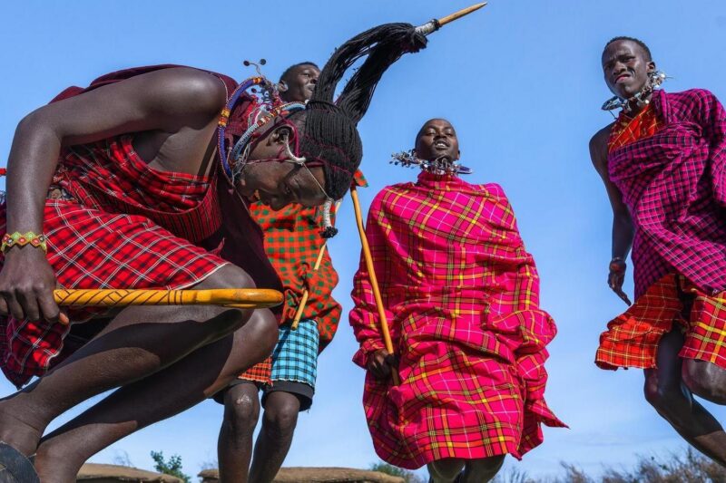 Maasai men in traditional red checkered clothing performing a high-jumping dance against a bright blue sky.