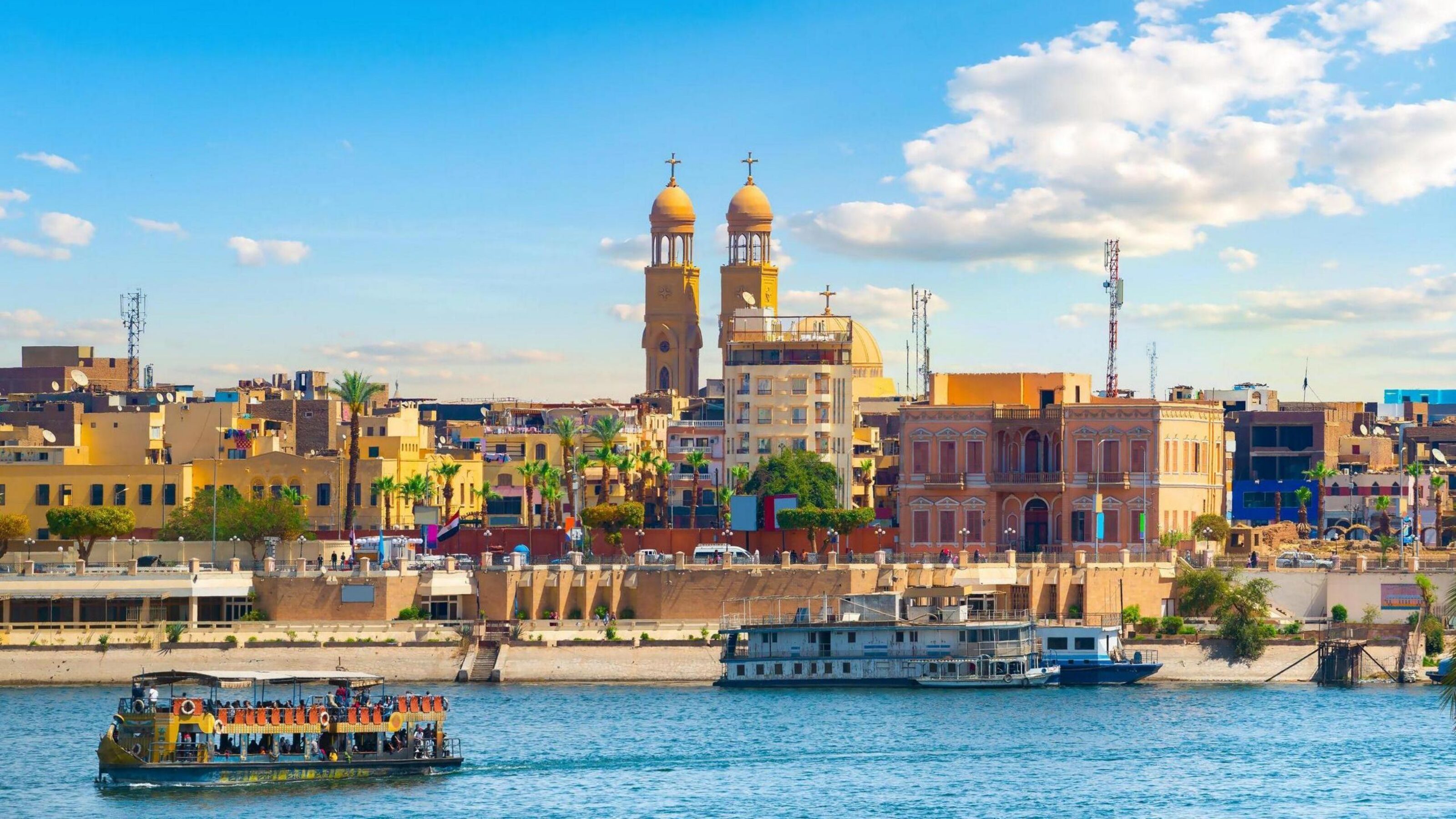 A scenic view of the Aswan skyline in Egypt featuring a large church with twin towers and boats on the Nile.