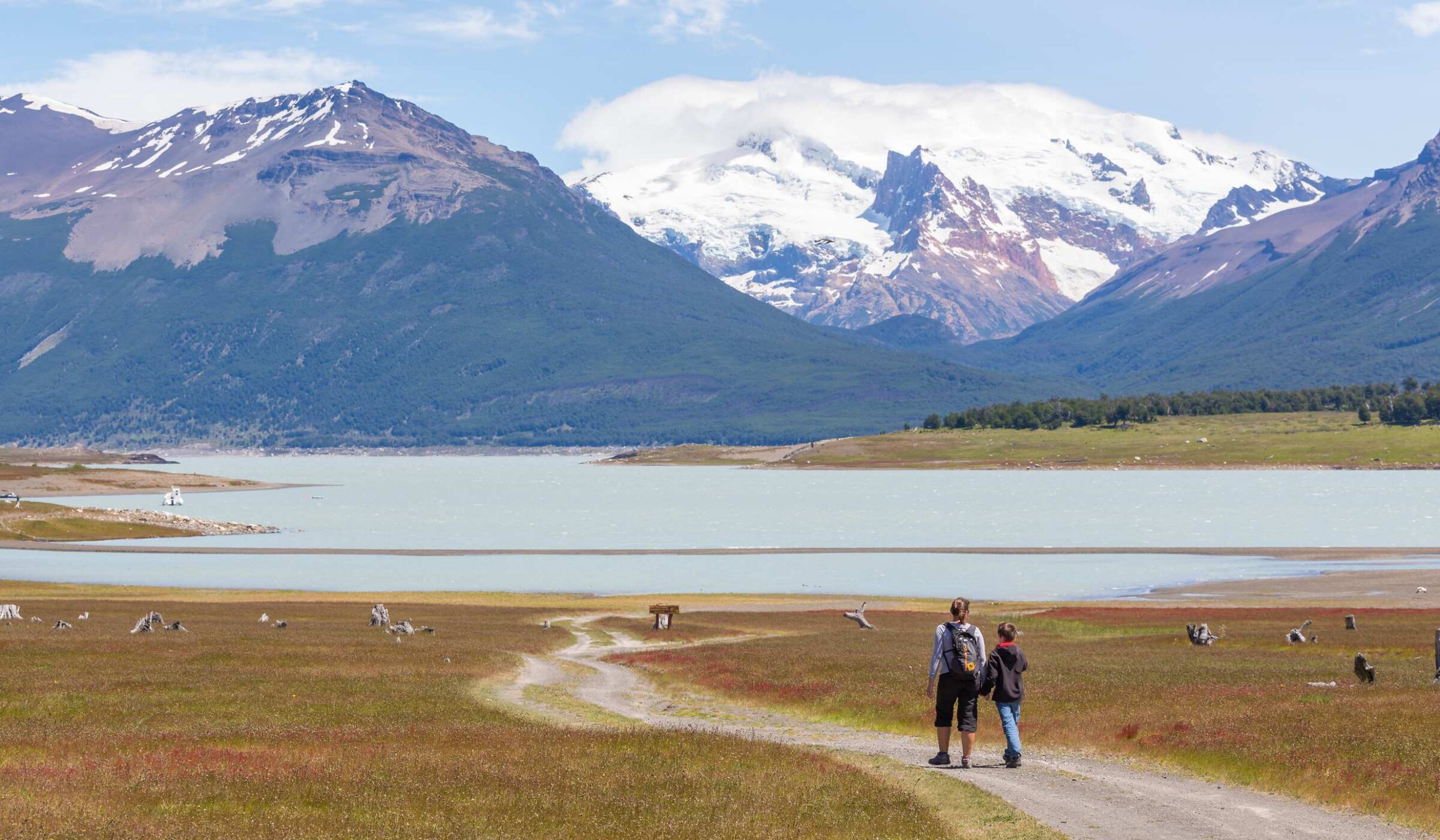 Two hikers walk down a dirt trail toward a large lake and snowy mountains in argentina patagonia.