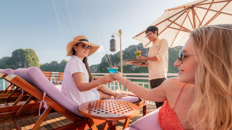 Two women toasting with blue and green cocktails on a sun deck overlooking the scenic islands of Halong Bay.