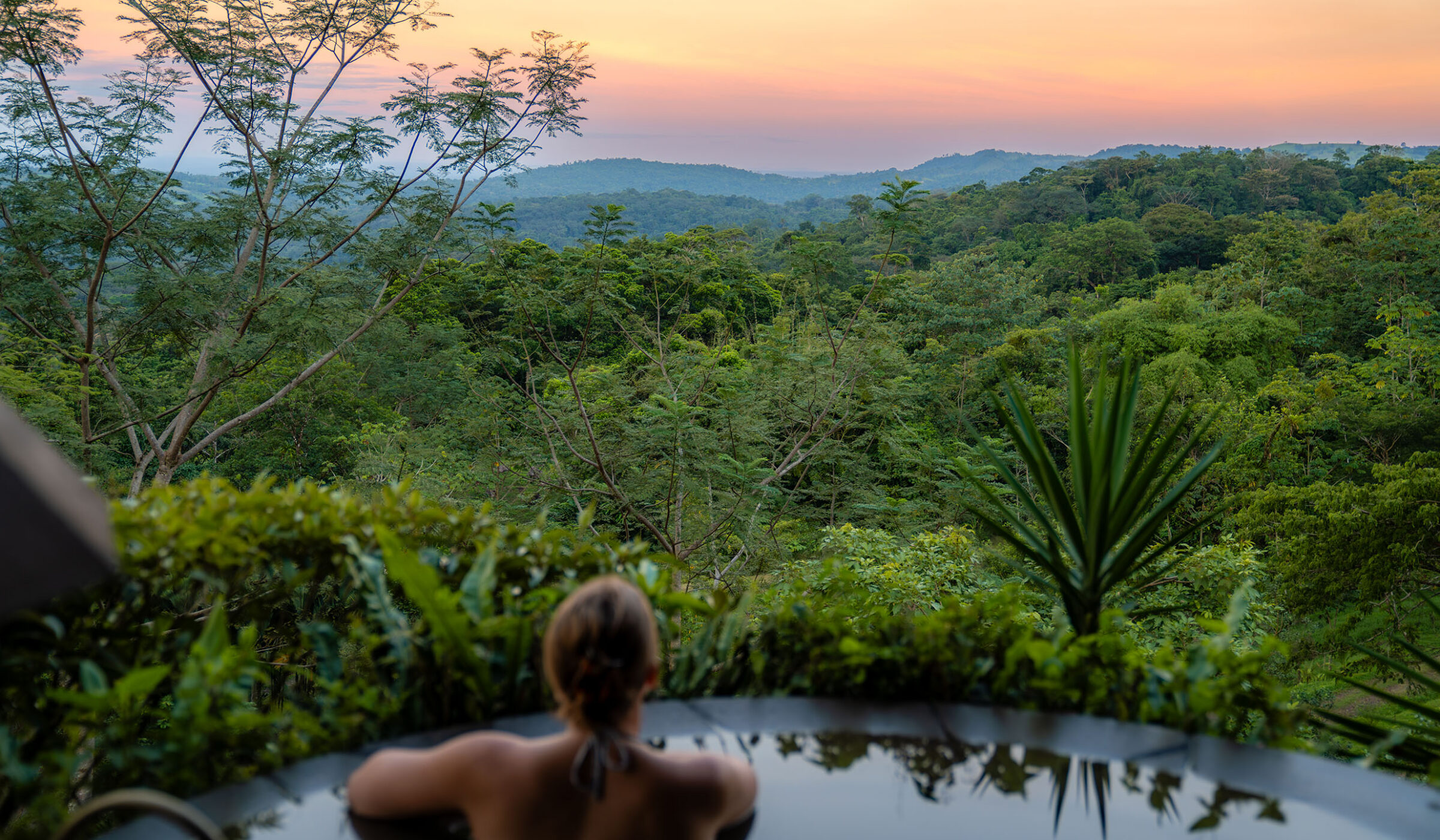 A woman in a plunge pool overlooking a vibrant jungle landscape as the sun sets in the horizon.