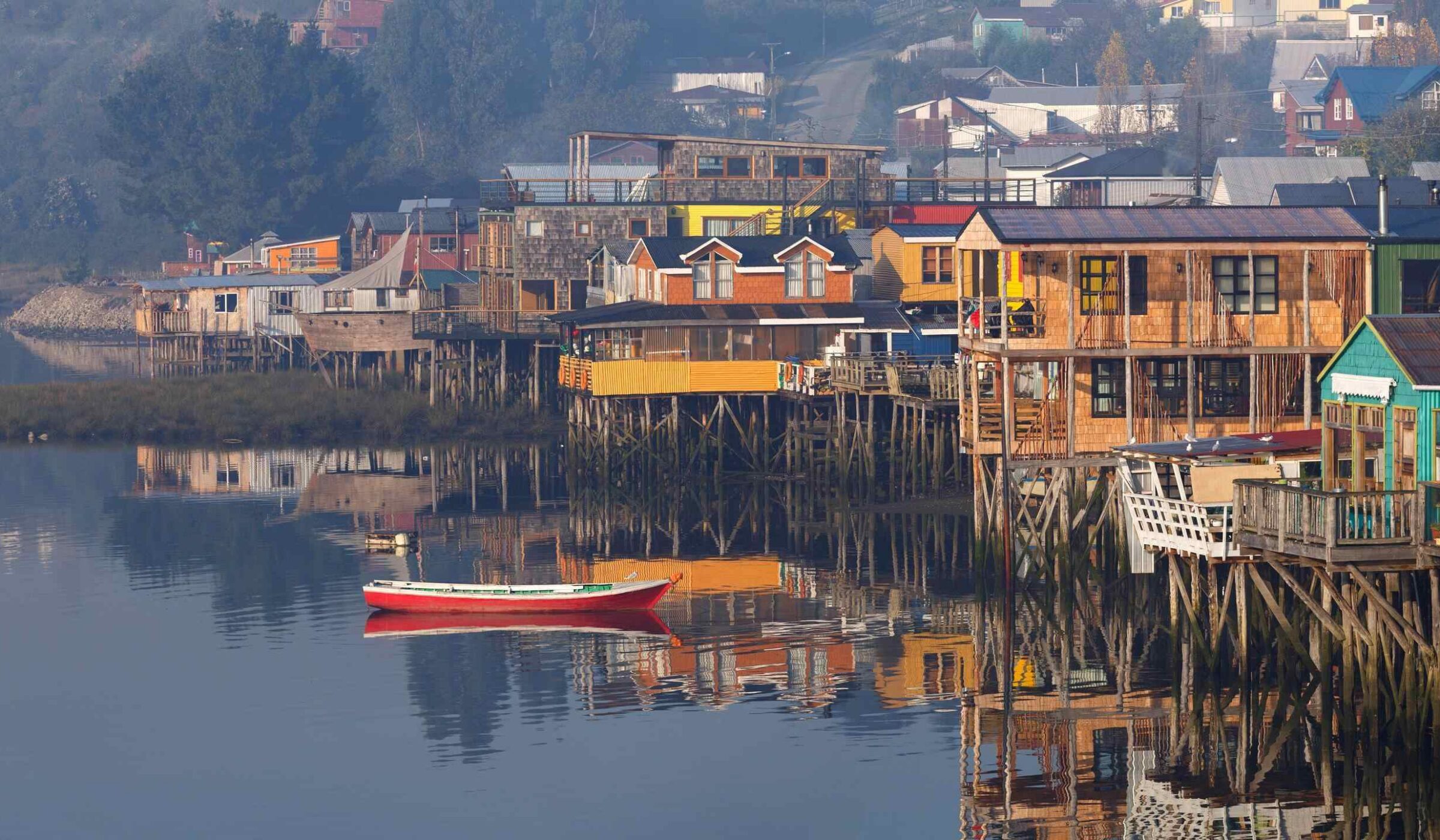 A red and white wooden boat on still water reflecting colorful houses built on stilts in the fog.