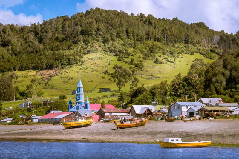 Fishing boats on a shore in front of a coastal village with a bright blue church and green hills.