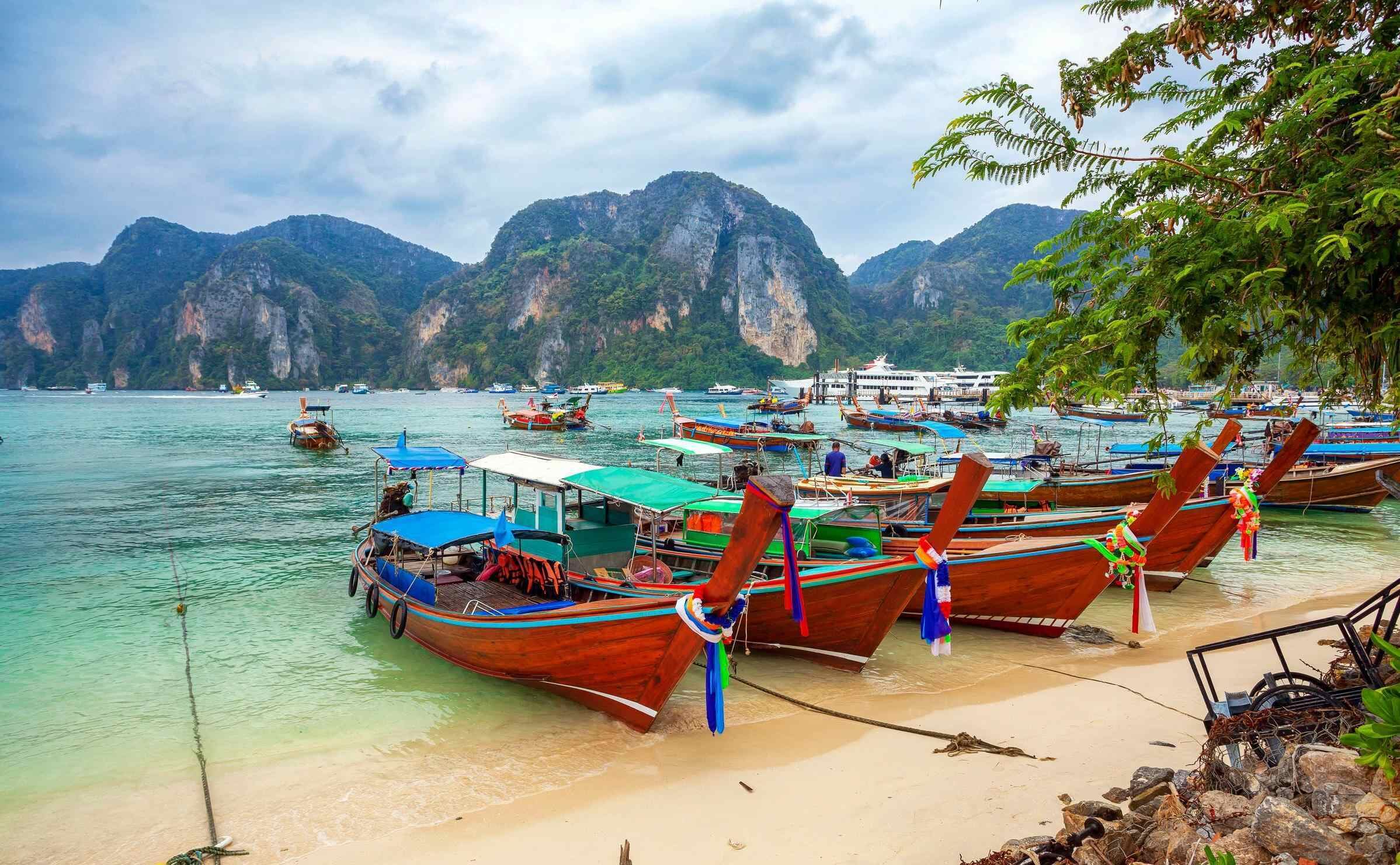 Longtail boats parked in a row on the beach with green sea and cliffs in the background in Phuket