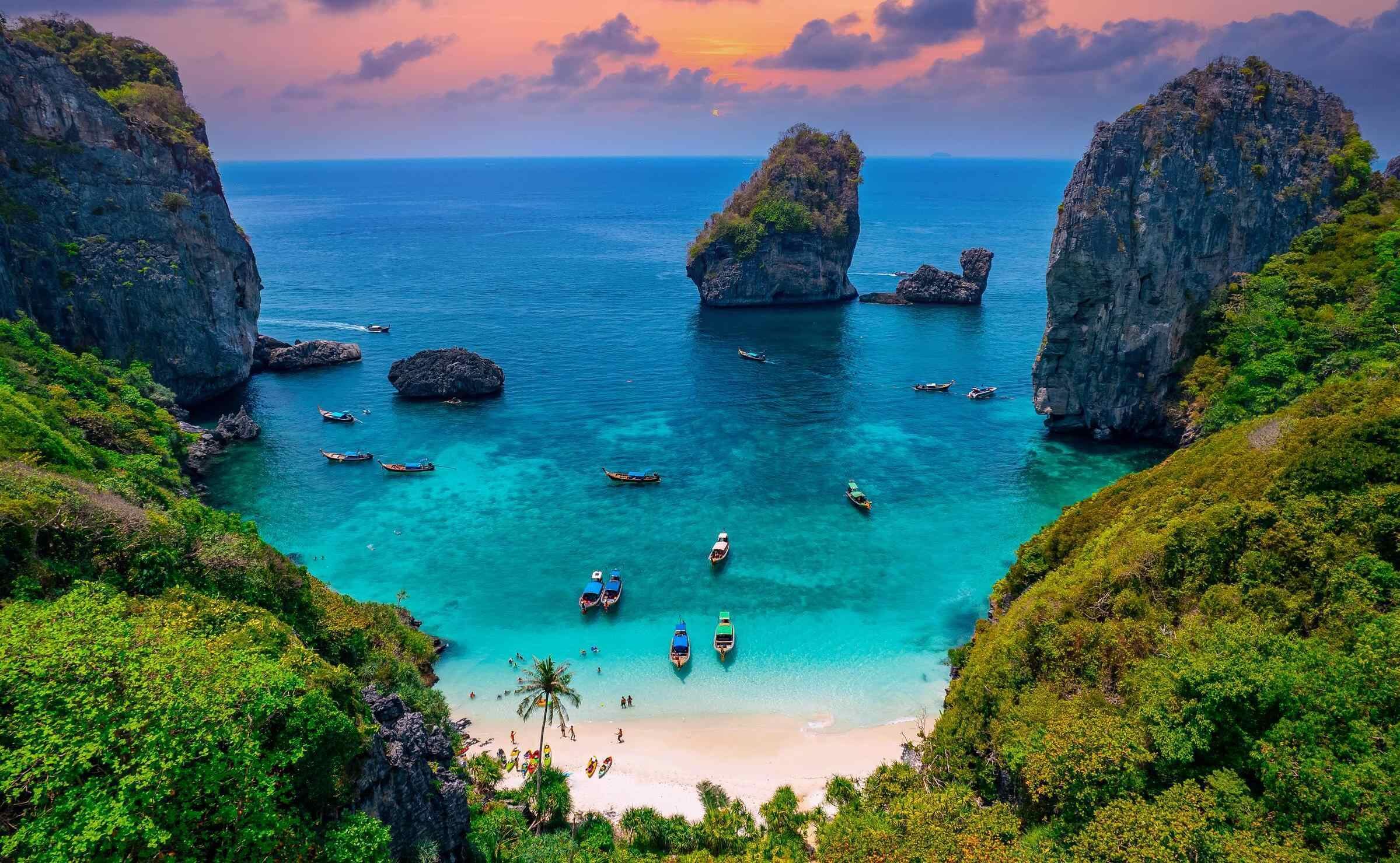 aerial view looking past tree covered cliffs onto a beach with turquoise sea and sunset sky
