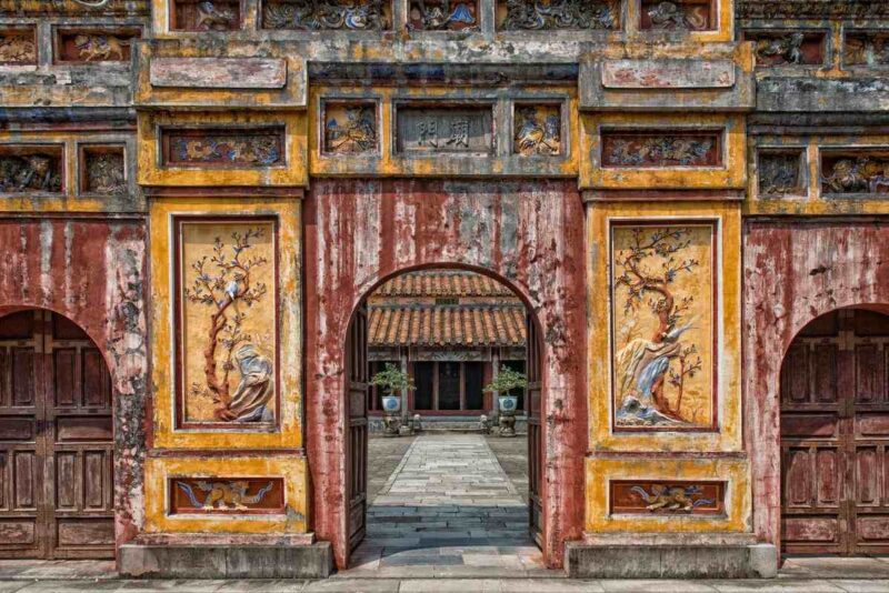 An ornate, weathered stone archway with decorative panels showing birds and trees leads into a courtyard with a temple.