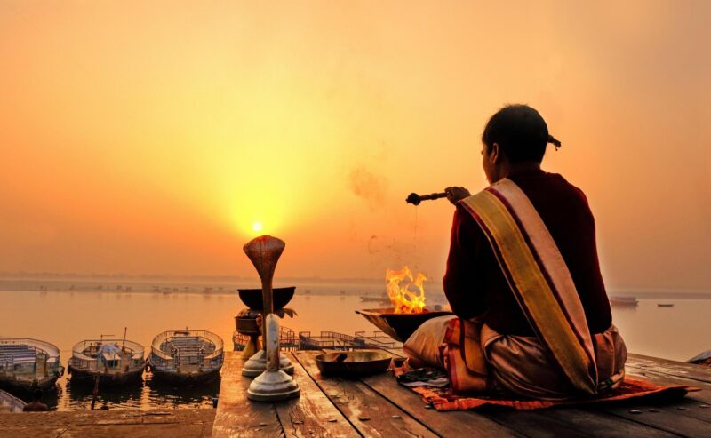 A person performing a ritual by a fire on a wooden dock at sunrise next to a calm river.