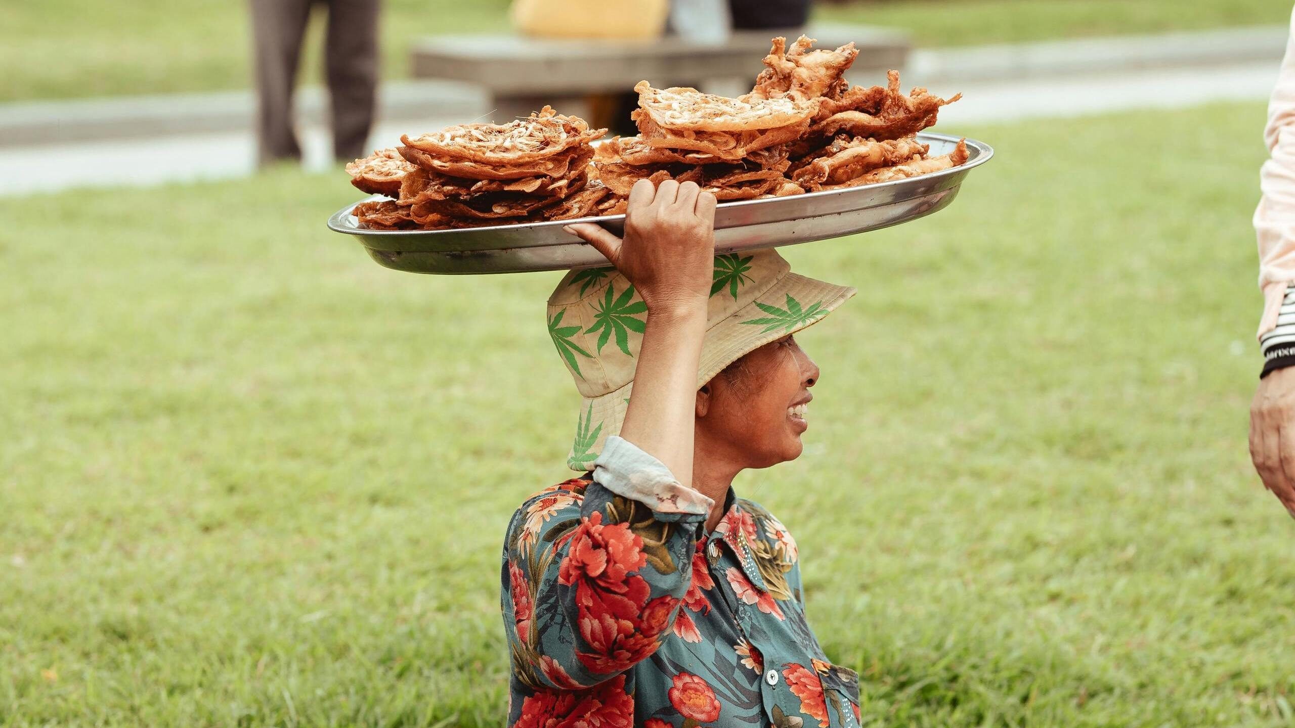 A person wearing a floral shirt and sun hat balancing a silver tray stacked with crispy fried snacks on their head.