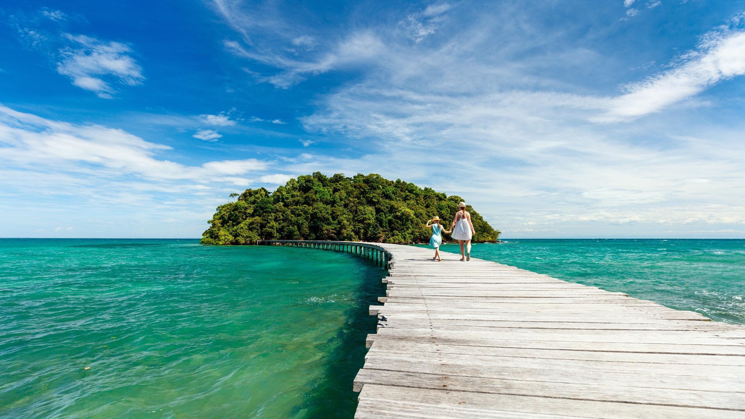 A woman and girl walk along a wooden bridge over blue ocean water toward a small forest-covered island.