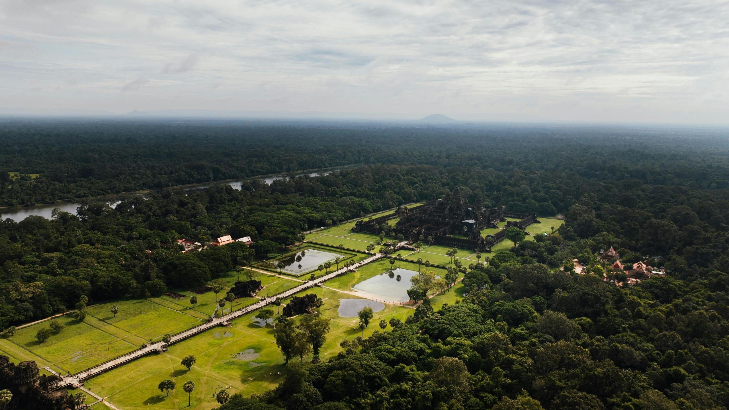 High-angle aerial view of ancient stone temple ruins and green lawns surrounded by a vast dense forest under a cloudy sky.