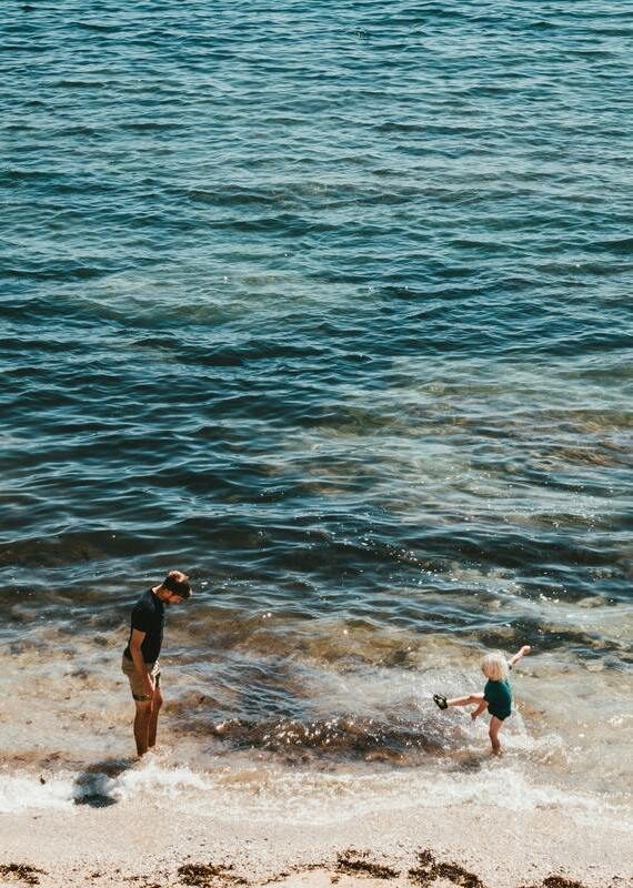 A man stands on a sandy shore watching a small child kick at the water's edge in the bright sunlight.