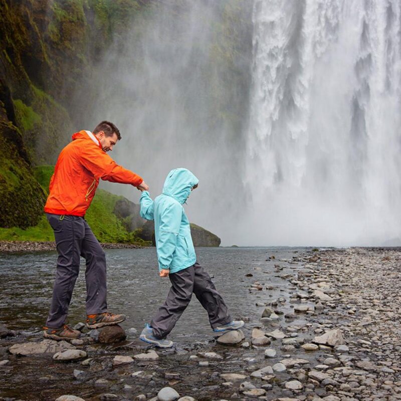 A father and child hiking across river stones at the base of a powerful waterfall on an Iceland family luxury tour.