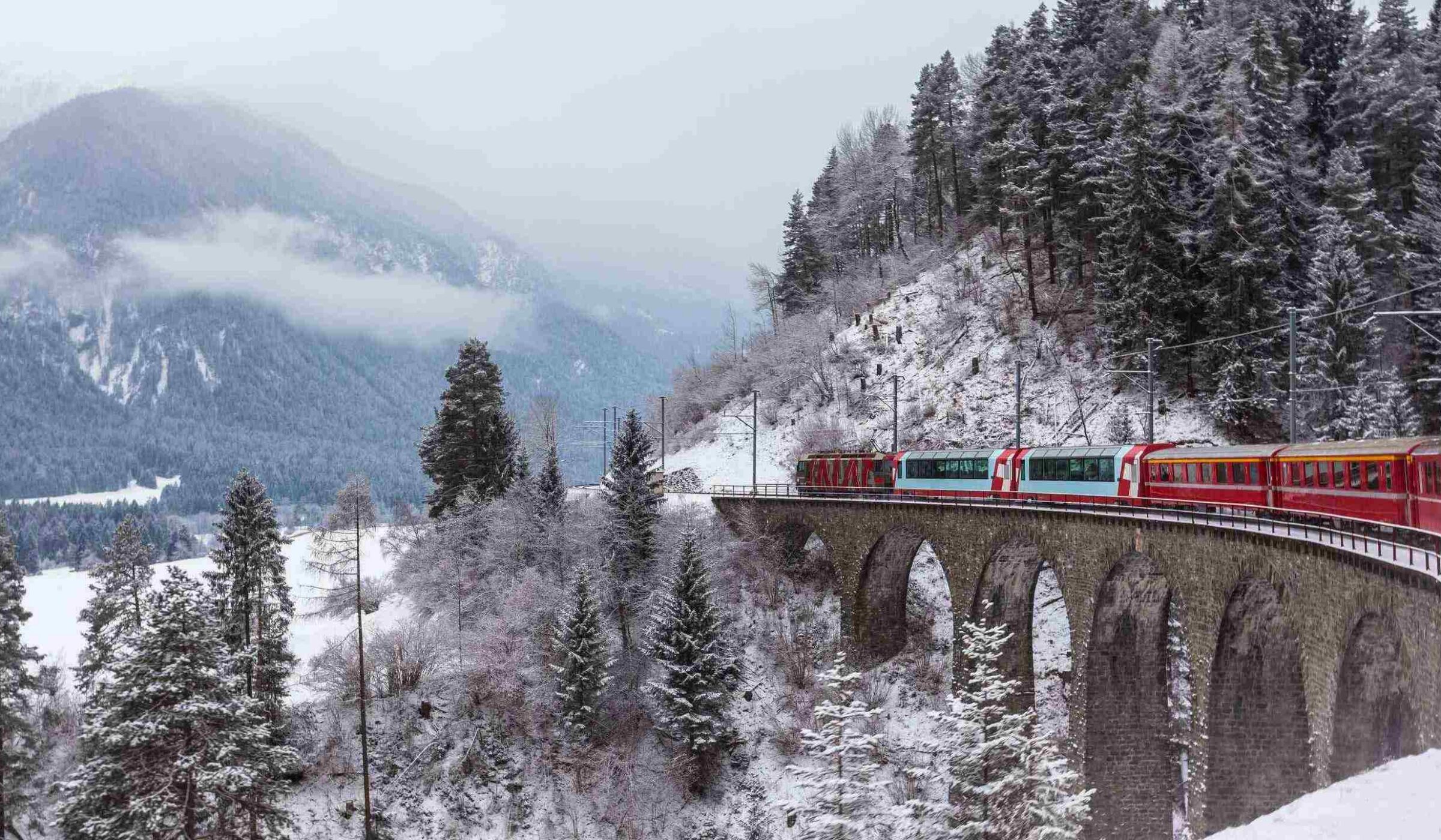 A red passenger train crosses a tall stone viaduct bridge in a snowy mountain forest in Switzerland.