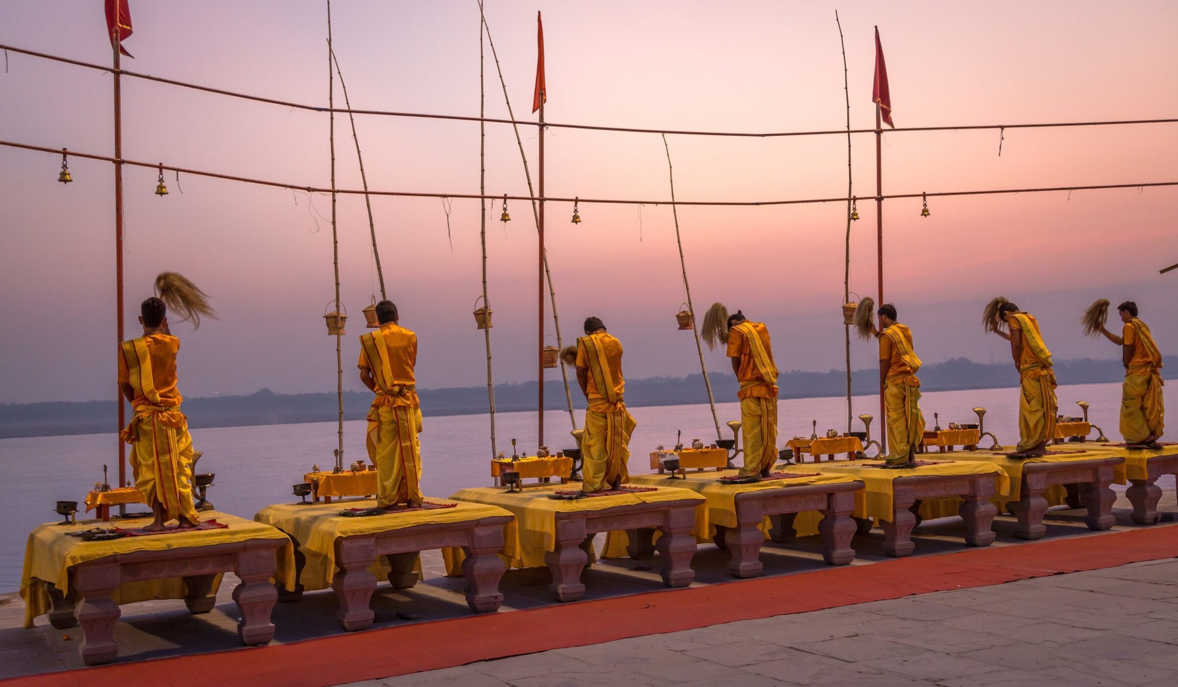 Priests in yellow robes perform a ritual with whisks on platforms overlooking a wide river during a pink sunset.