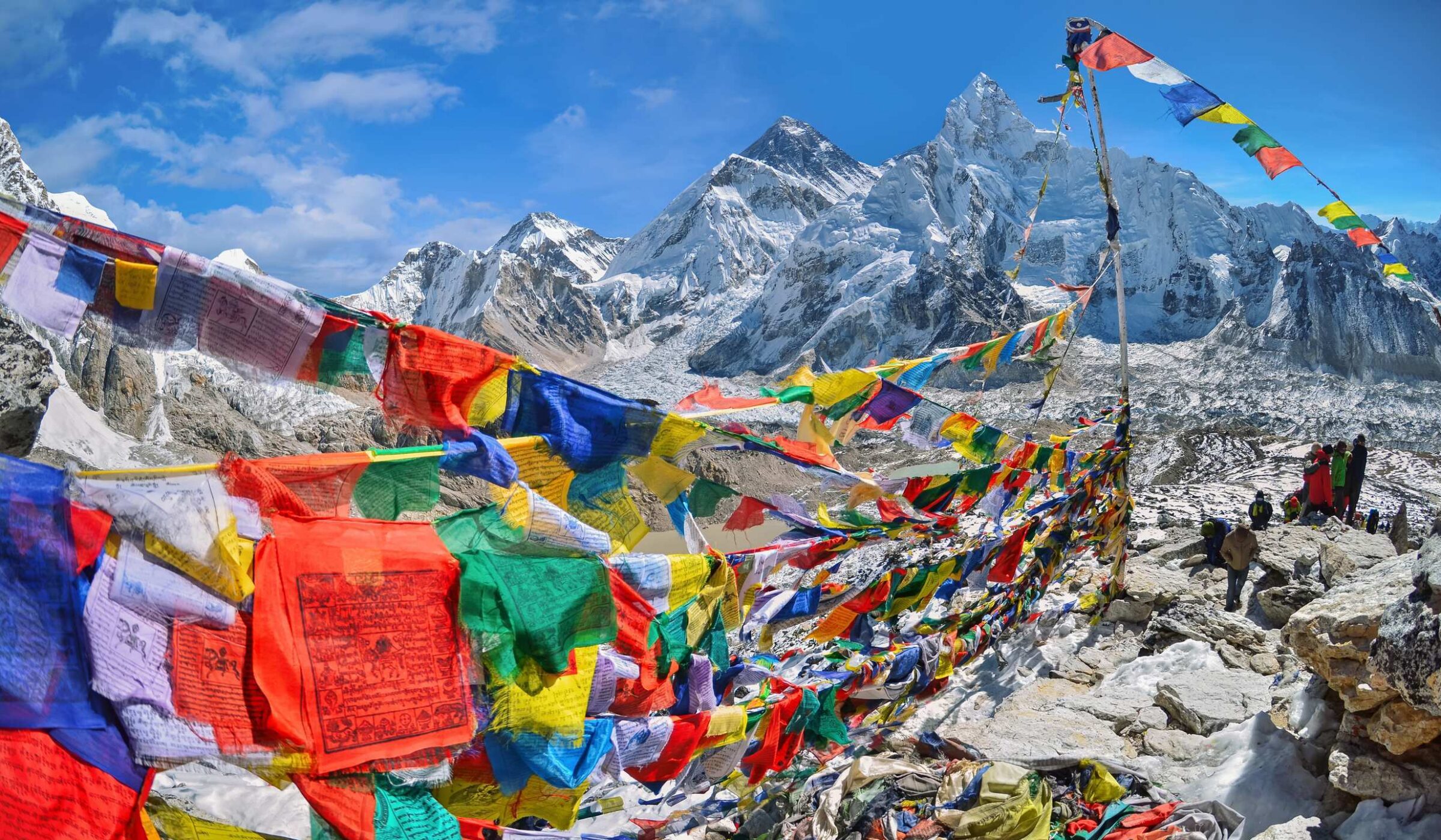 Colorful prayer flags stretch across a rocky mountain pass with massive snow-covered Himalayan peaks in the background.
