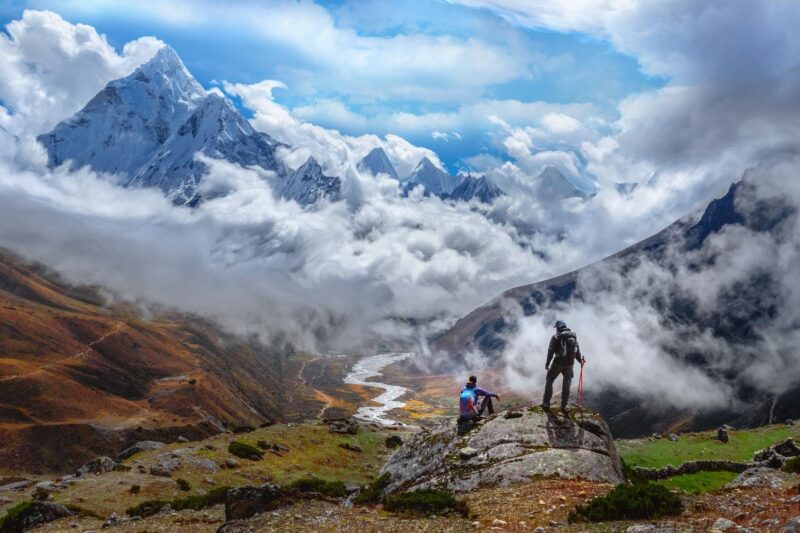 Two trekkers stand on a rock overlooking a deep valley with snow-capped mountains peaking through thick white clouds.