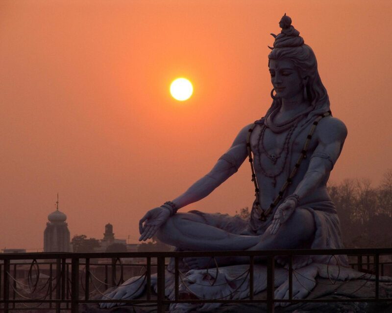 A large statue of Lord Shiva in a seated meditation pose against a glowing orange sunset sky and distant temple.