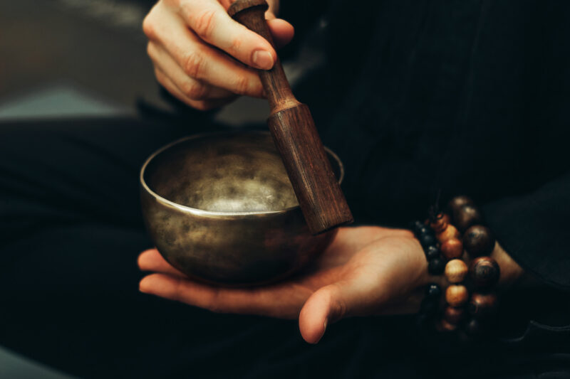 Close-up of a person holding a bronze singing bowl and wooden mallet, wearing beaded bracelets during a sound bath.