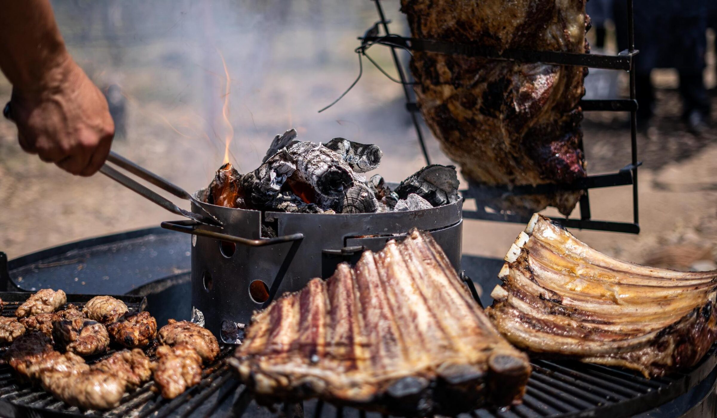 Beef ribs and assorted meats grilling over hot coals and flames on a large outdoor metal barbecue.