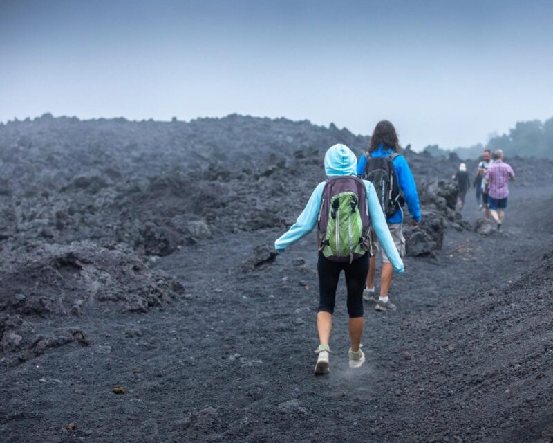 Two hikers with backpacks walk on a trail made of dark volcanic rock toward other people in the distance.