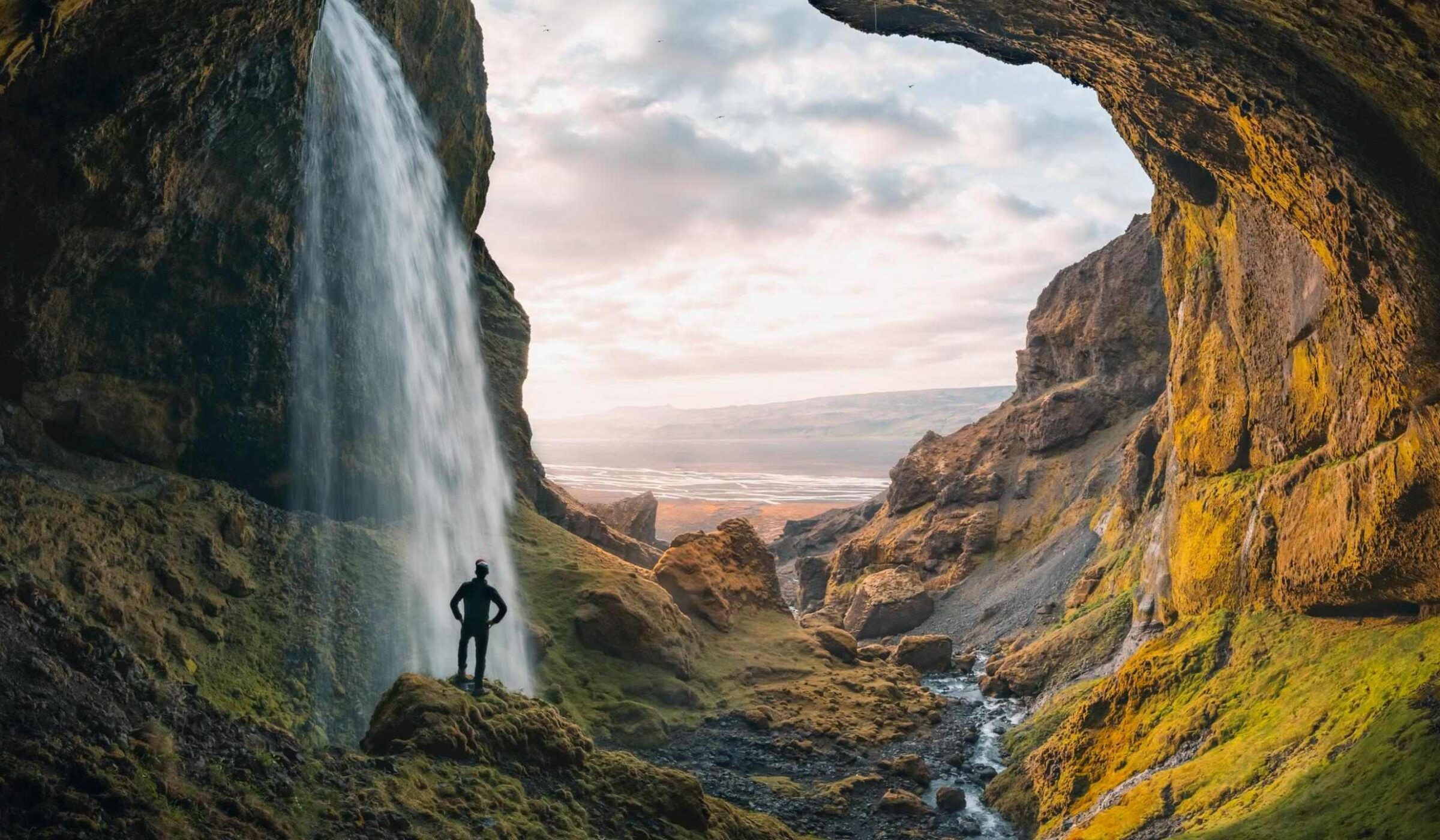Silhouette of a person standing by a large waterfall flowing from a rocky cave opening overlooking a distant valley.