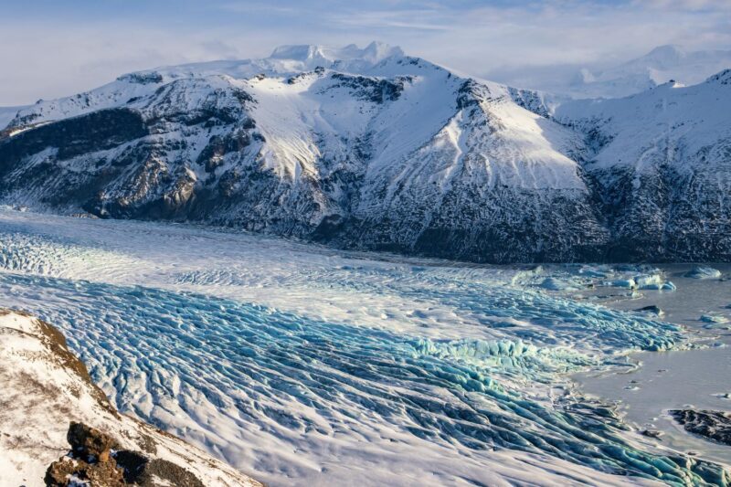 An expansive glacier with blue and white ice ridges sits at the base of snow-covered mountains in Iceland.