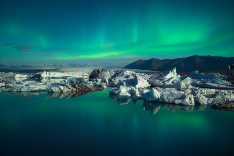 Green northern lights illuminate the night sky above a lagoon with icebergs and distant mountains in Iceland.