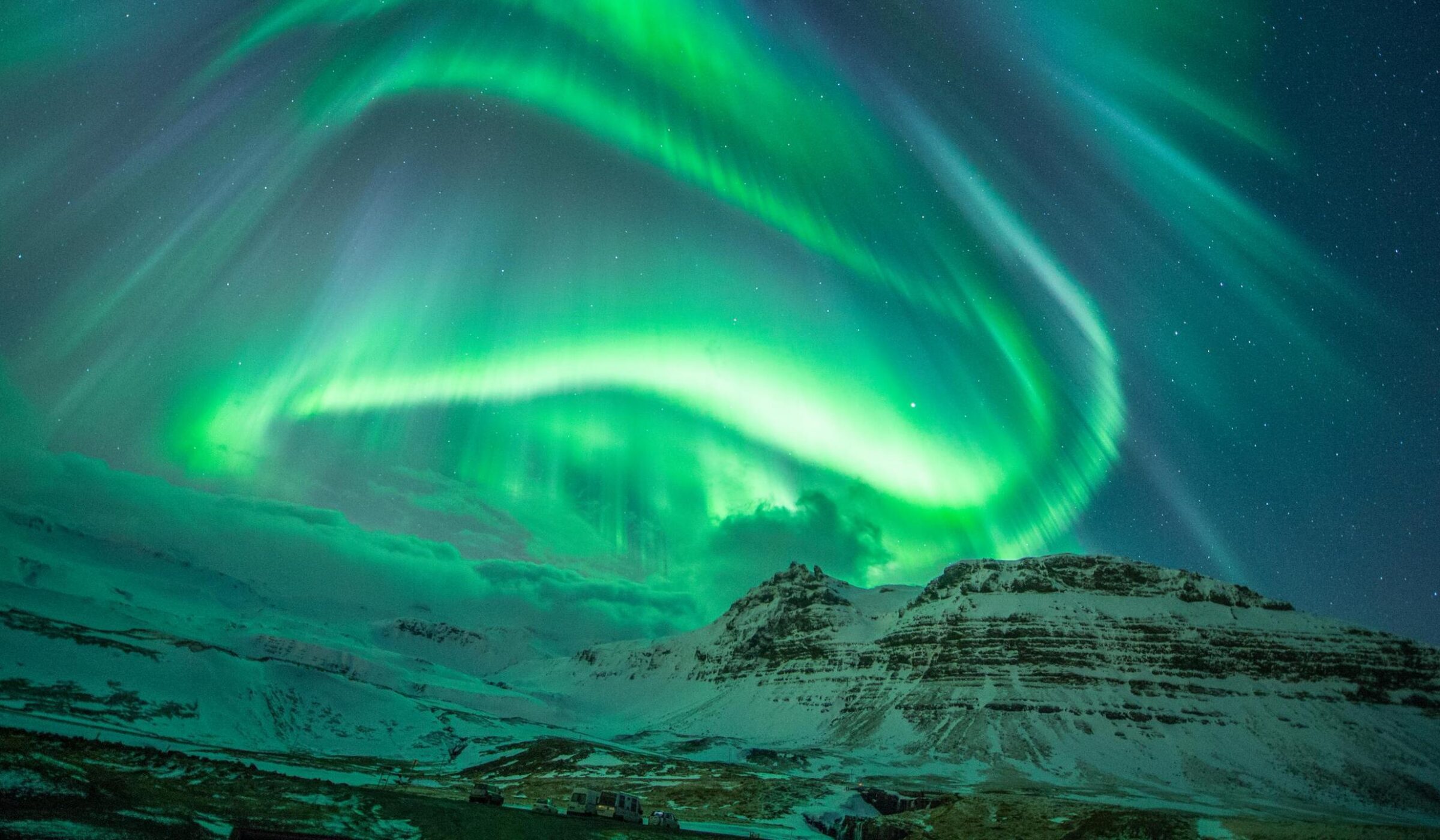 Swirling green aurora borealis lights dance in the night sky over a snowy mountain landscape in Iceland.