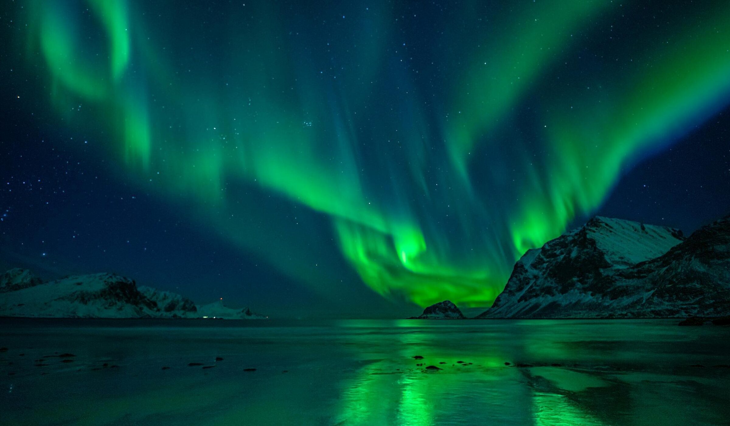 Green northern lights illuminate a starry night sky above snowy mountains and a reflective body of water.