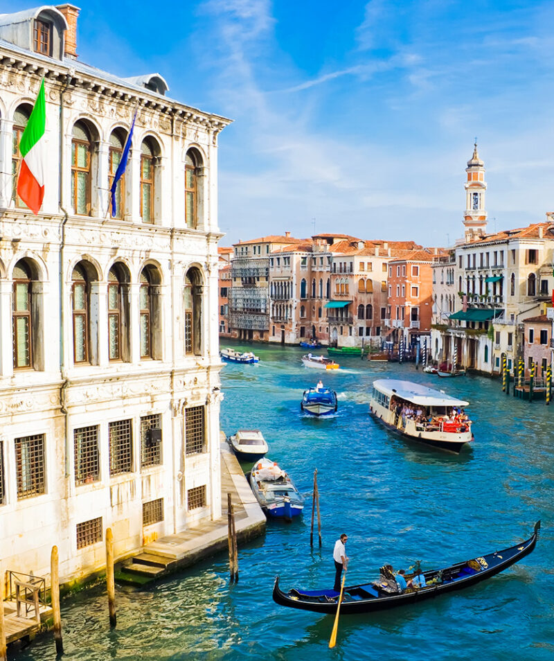 A gondolier navigates a black gondola on a busy blue canal in Venice, lined with historic Venetian buildings.