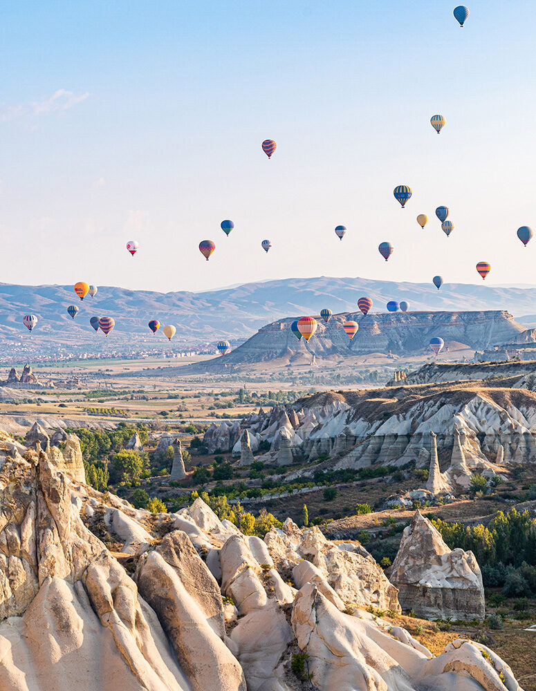Dozens of colorful hot air balloons fly over rocky fairy chimney formations and rolling hills under a clear blue sky.