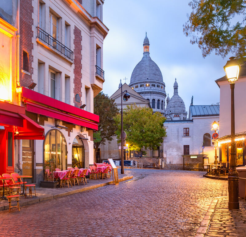 A Parisian street cafe with red chairs and tables on a cobblestone road, with a large domed basilica in the background.