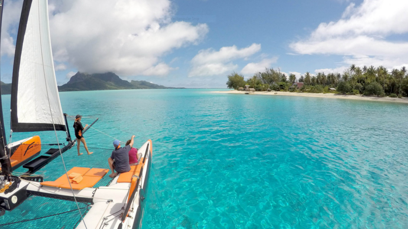 People on a catamaran sailing through clear turquoise water toward a tropical island coastline.