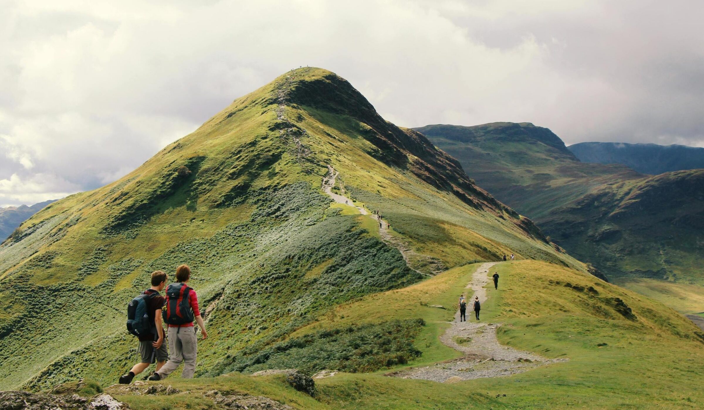 Two hikers with backpacks walking along a mountain ridge path leading toward a sharp green peak.