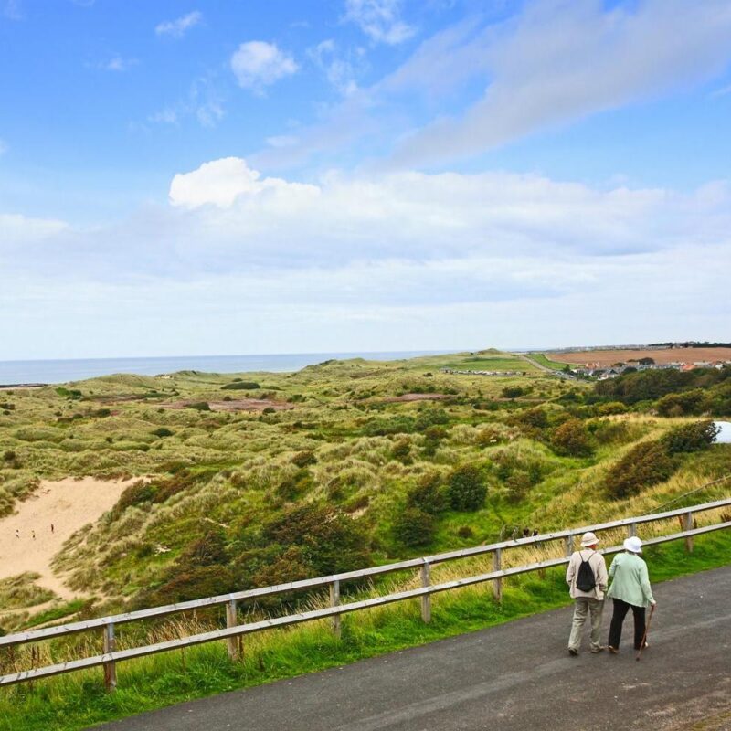 Older couple walking on a paved coastal path next to rolling green sand dunes and the distant sea.