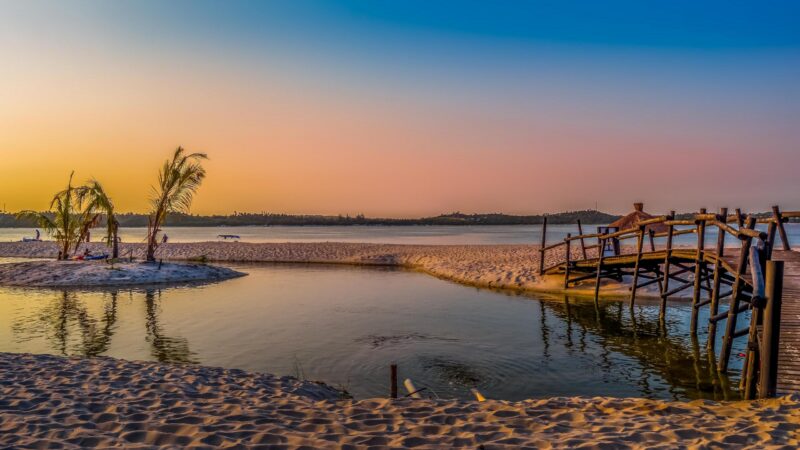 A wooden footbridge next to a beach lagoon with palm trees under a vibrant orange and blue gradient sunset sky.