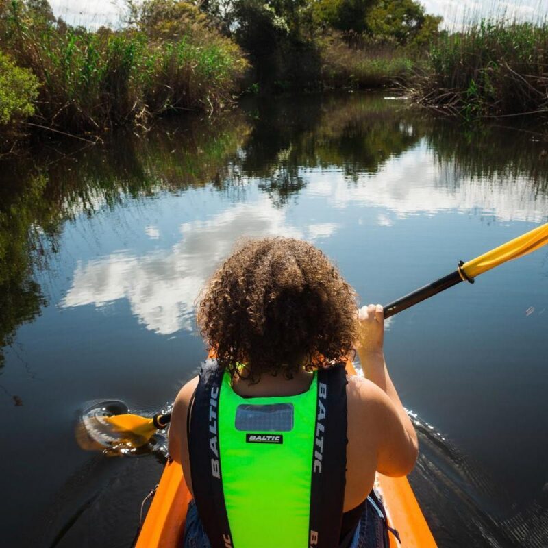 First-person rear view of a person with curly hair paddling a canoe down a still waterway under a bright sky.