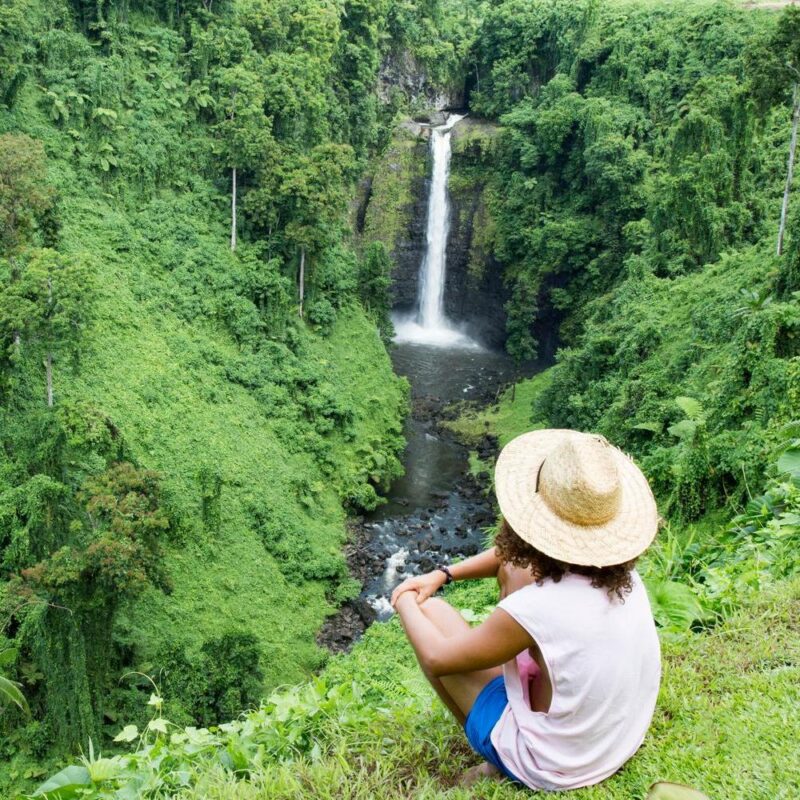A person in a straw hat sits on a grassy cliff looking at a tall waterfall in a lush green valley.
