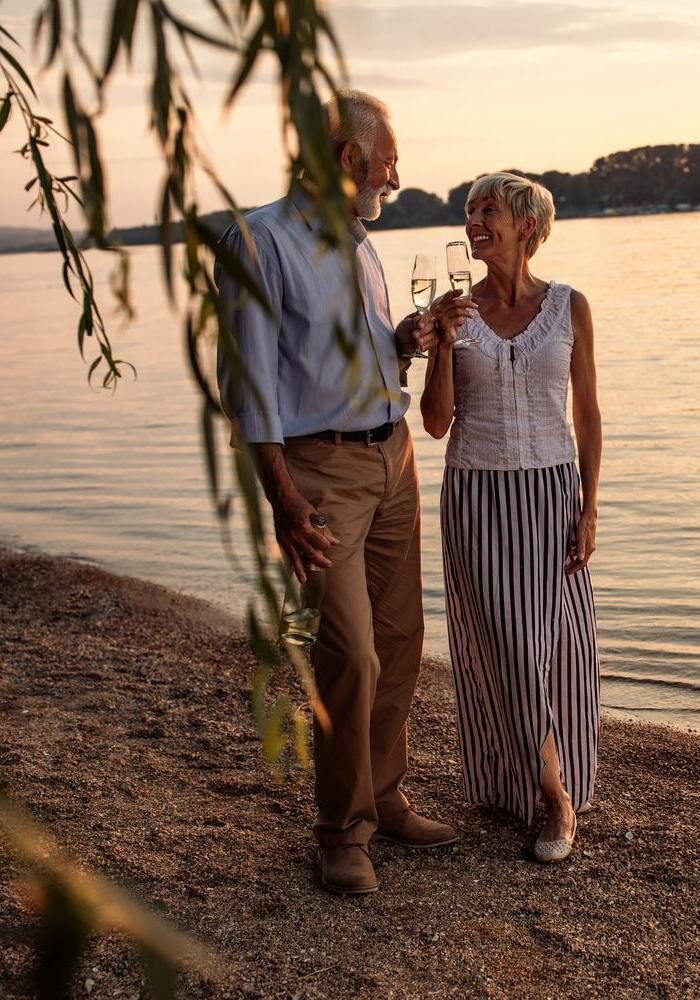 A couple holding wine glasses and smiling at each other on a beach at sunset.