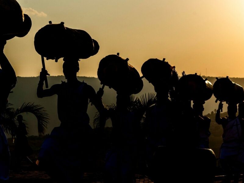 Silhouettes of dancers carrying large drums on their heads against a bright sunset sky.