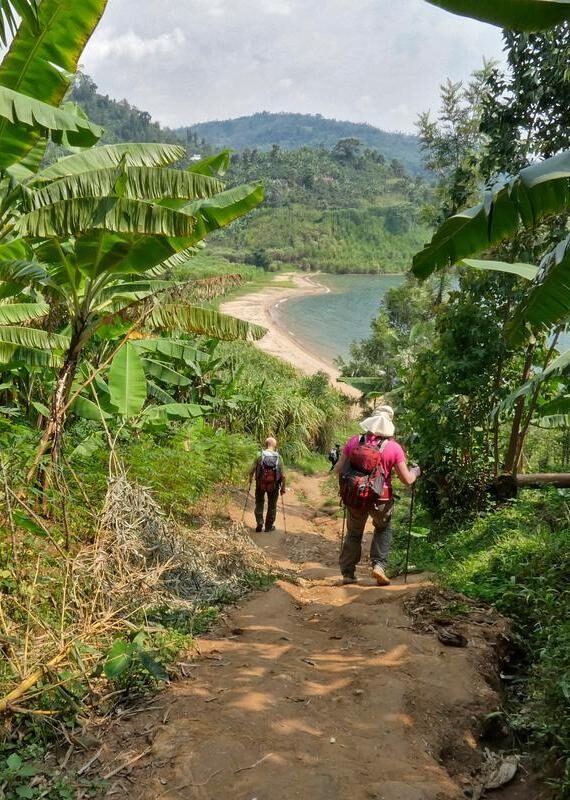 Two people hiking a dirt path surrounded by lush banana trees leading to a lakeside beach.