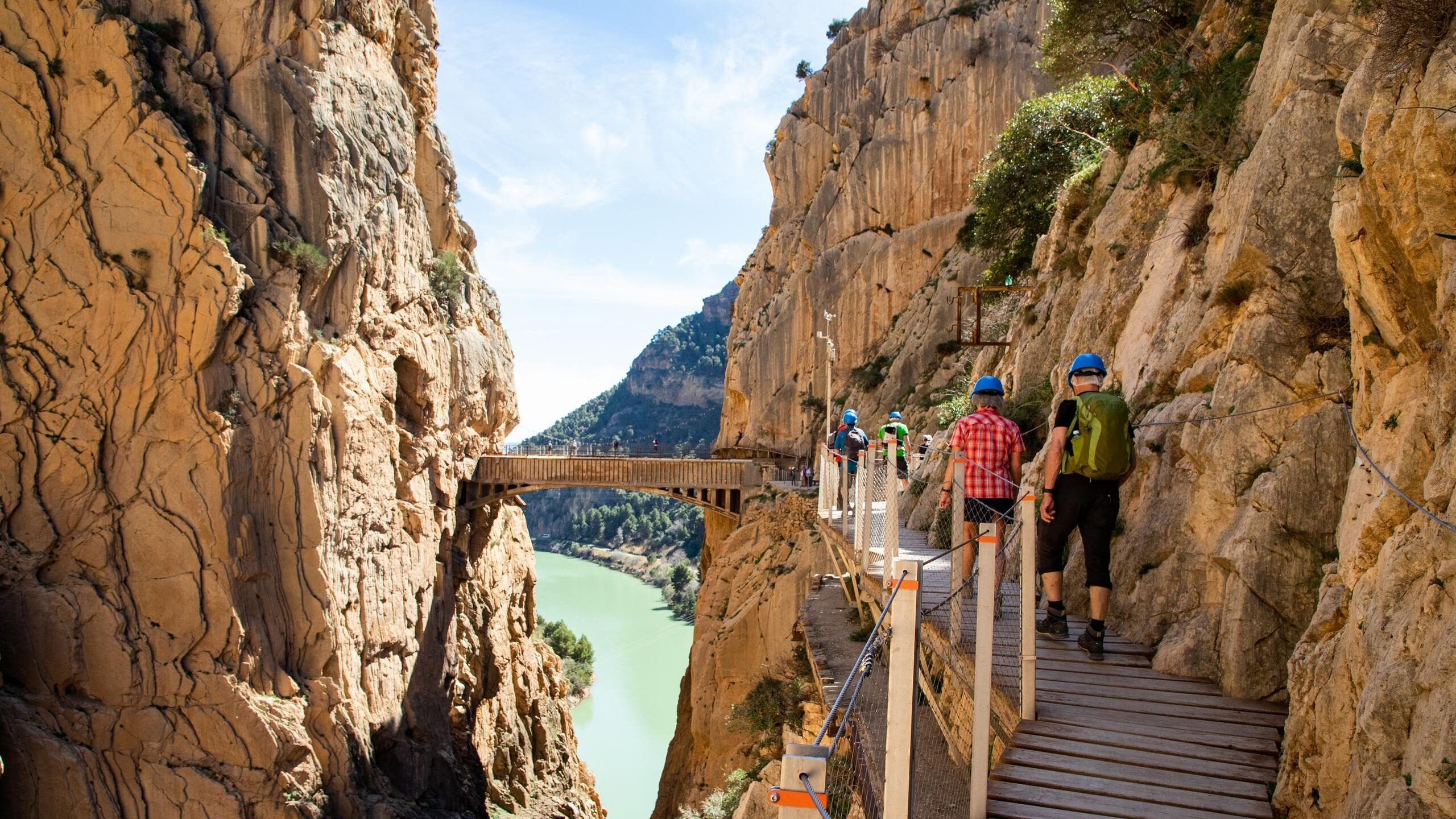 Hikers in helmets walk along a narrow wooden boardwalk attached to a steep rocky cliff over a river at Caminito del Rey.