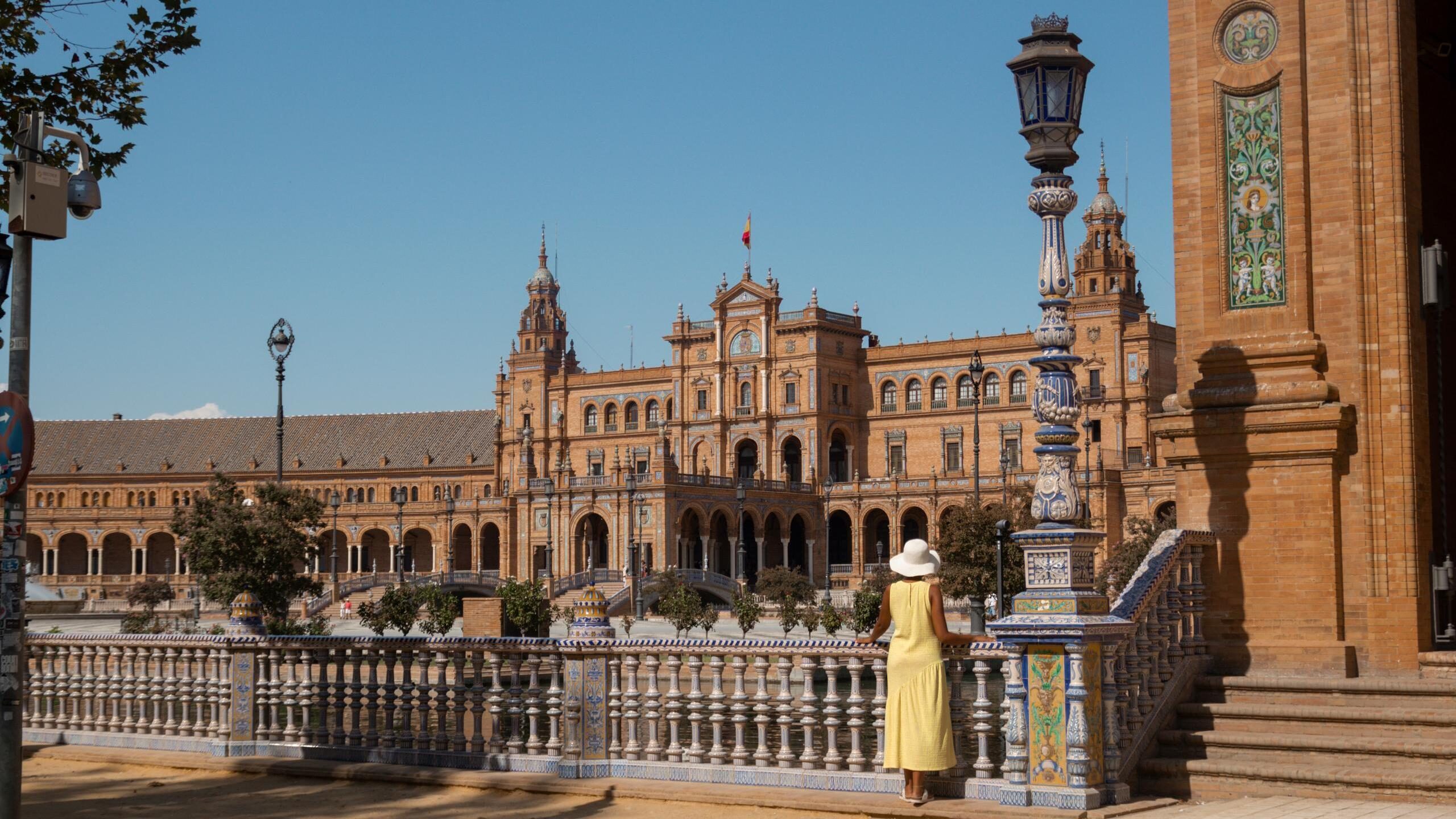 A woman in a yellow dress and hat stands by an ornate tiled railing at Plaza de España in Seville.