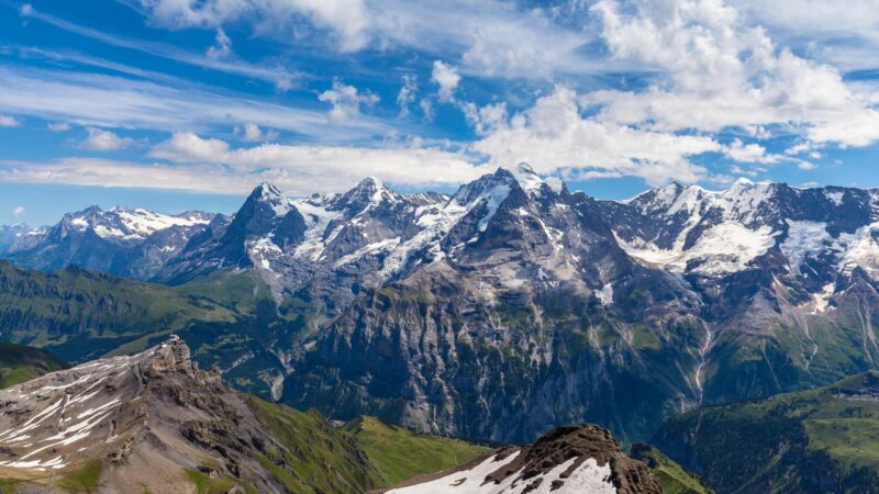 Panoramic view of a rugged, snow-capped Swiss mountain range rising above deep green valleys under a cloudy blue sky.